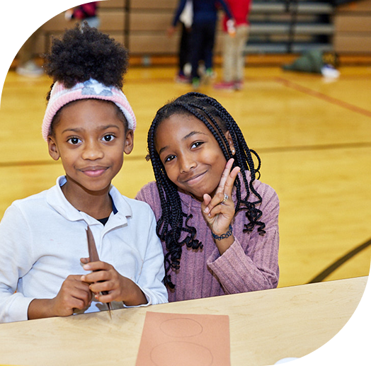 Two children smiling and sitting at a table in a gymnasium.