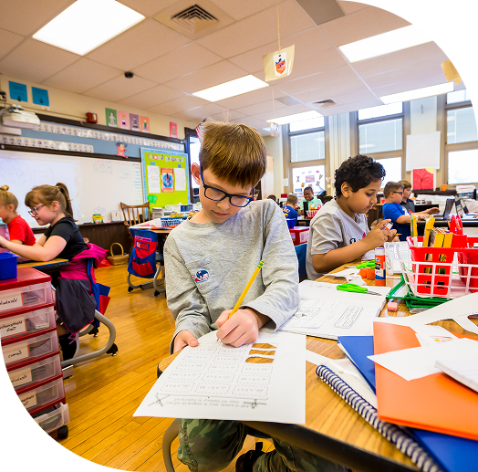 A classroom filled with students engaged in various activities, with one child focused on writing at their desk.