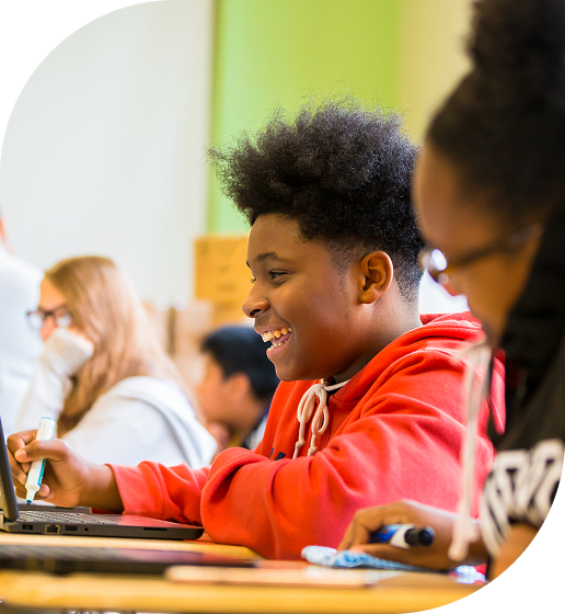 A student in a red hoodie is smiling while using a laptop in a classroom setting.