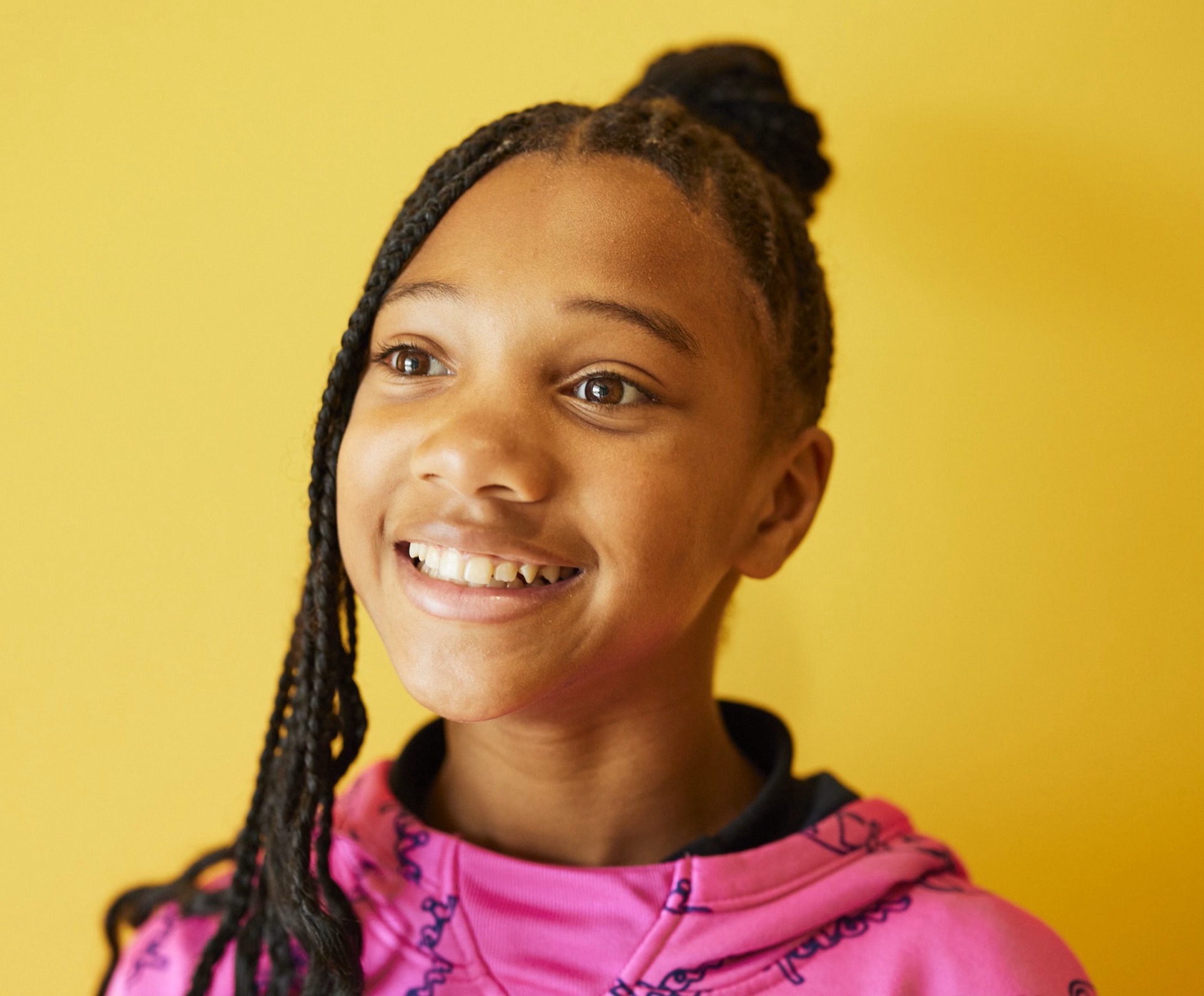 A person with braided hair smiles against a bright yellow background while wearing a pink top.